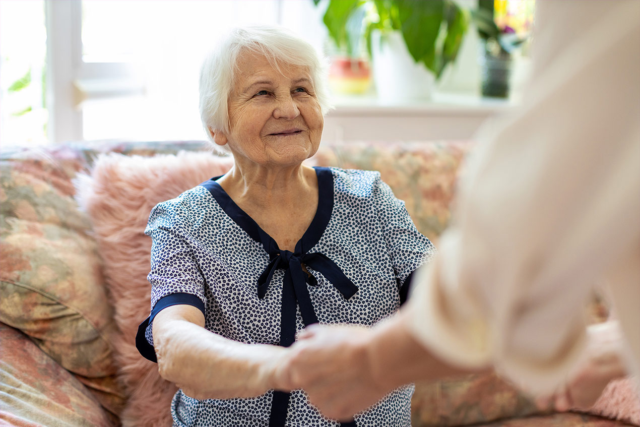 Eine Seniorin sitzt auf einem Sofa und wird von einer Person an den Händen gehalten