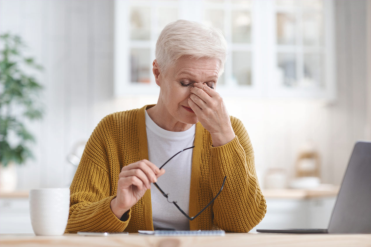 Eine Frau fasst sich an den oberen Nasenrücken und hällt in der rechten Hand ihre Brille.