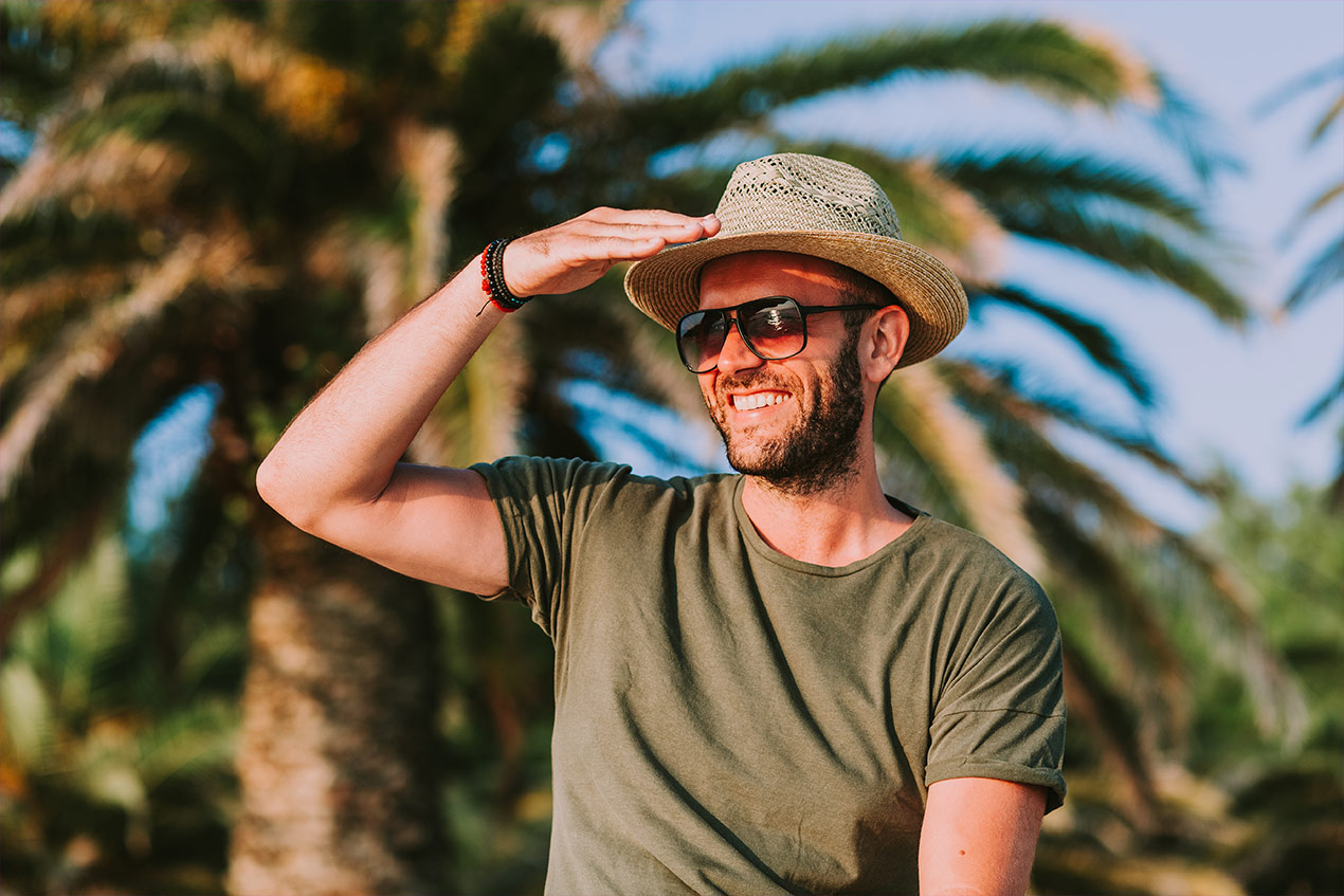 Mann im Urlaub mit Sonnenbrille und Hut steht vor Palmen