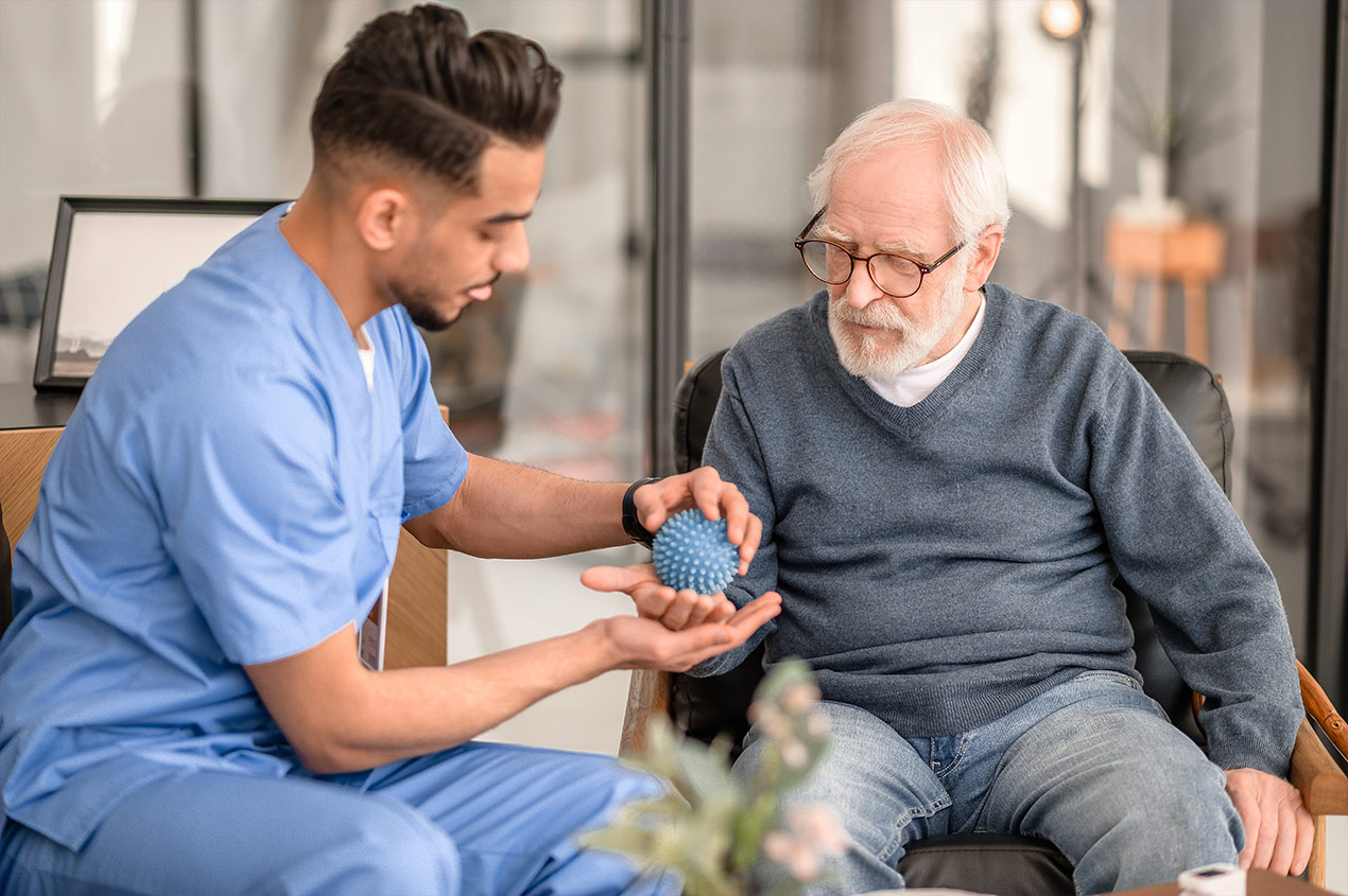 Ein Pfleger führt bei einem Senior eine Behandlung der Hand mit einem Igelball durch