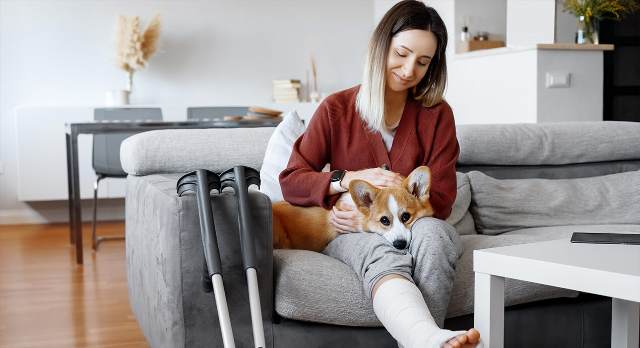 Frau mit Gipsbein, die Krankengeld erhält, sitzt auf einer Couch und streichelt einen Hund auf Ihrem Schoß