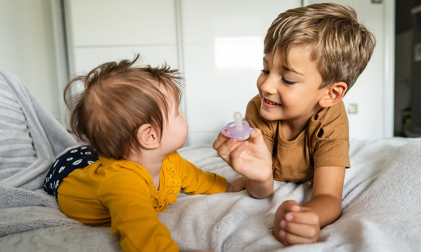 Zwei kleine Geschwisterkinder, der große Bruder hält dem kleineren Geschwisterkind den Schnuller hin